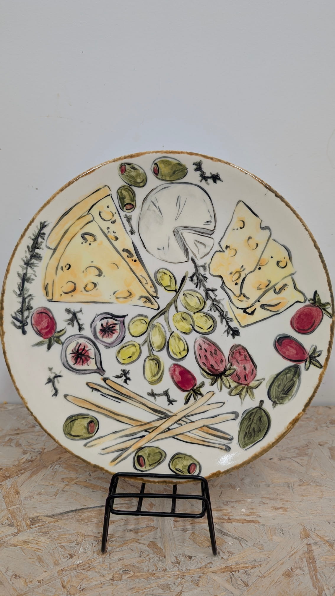 Ceramiczne walentynki 3 Biały, ceramiczny talerz z ręcznie malowaną ilustracją deski serów i przekąsek. Na środku widać kiść zielonych winogron, wokół której rozmieszczono kawałki żółtego sera z dziurami, krążek camemberta, przekrojone figi, truskawki, oliwki oraz paluszki chlebowe. Całość wykonana w radosnym, lekko ilustracyjnym stylu z wyraźnym, czarnym konturem.