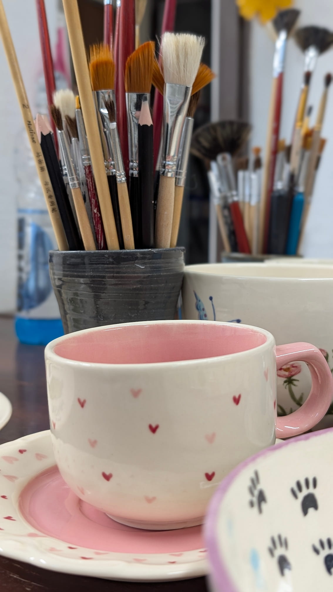 Ceramiczne walentynki 4 Biała filiżanka z jasnoróżowym uchem i różowym wnętrzem, stojąca na pasującym spodku. Zewnętrzna strona ozdobiona jest regularnym wzorem drobnych, czerwonych i różowych serduszek. W tle widoczne są pędzle malarskie w pojemniku, sugerujące pracownię ceramiczną.