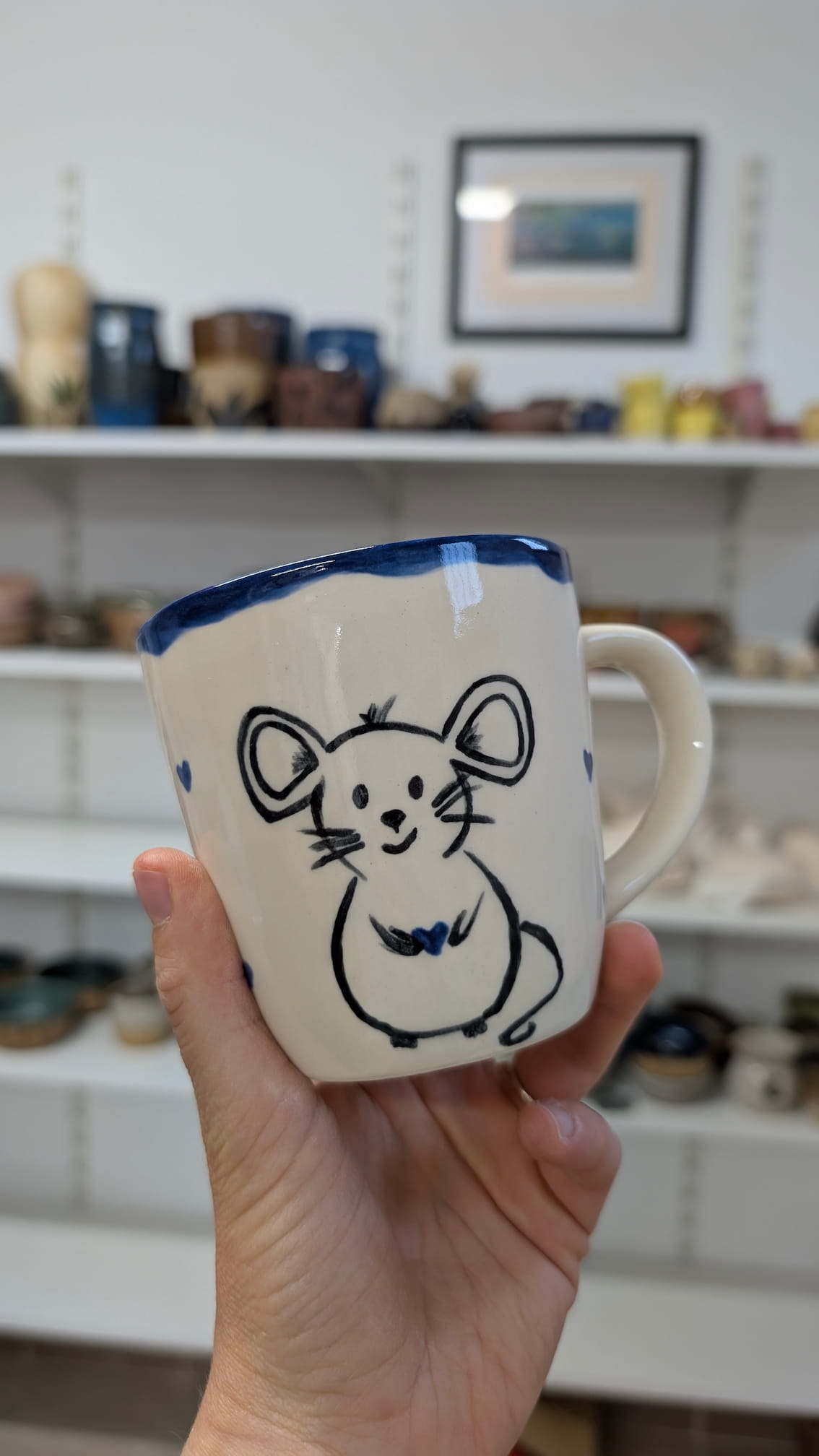 Ceramiczne walentynki 5 Ręcznie malowany kubek z białą glazurą. Na froncie widnieje urocza, czarna ilustracja myszki trzymającej małe, niebieskie serduszko. Górna krawędź kubka jest nieregularnie pomalowana ciemnoniebieską farbą, a po bokach widać pojedyncze, małe serca.
