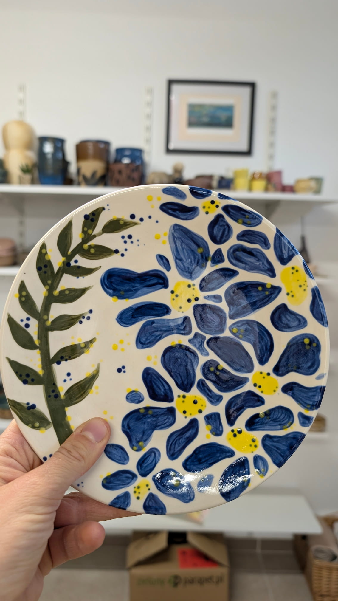 Ceramiczne walentynki 10 Ceramiczny talerz ozdobiony ekspresyjnym motywem roślinnym. Większą część zajmują duże, ciemnoniebieskie płatki kwiatów z żółtymi środkami. Po lewej stronie widnieje długa, ciemnozielona gałązka z listkami. Tło uzupełniają drobne, żółte i niebieskie kropki, nadając całości dynamiczny charakter.