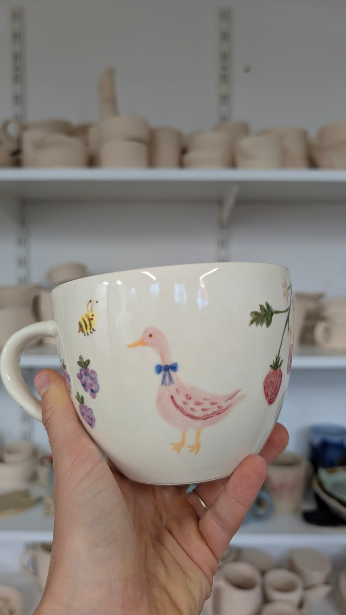 Ceramiczne walentynki 8 Duży, pękaty kubek w kolorze kremowym. Głównym motywem jest ilustracja różowej kaczki z niebieską kokardą na szyi. Obok kaczki namalowano małą pszczółkę, fioletowe owoce (jagody) oraz truskawkę zwisającą z gałązki. Styl malunku jest delikatny i sielankowy.