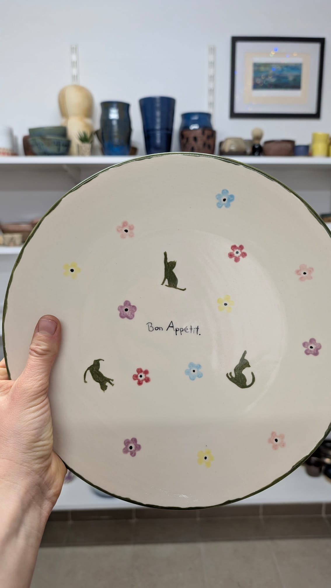 Ceramiczne walentynki 7 Minimalistyczny talerz z kremowym tłem. Na środku widnieje czarny, odręczny napis „Bon Appétit”. Wokół napisu rozproszone są małe, kolorowe kwiatuszki (różowe, niebieskie, żółte, fioletowe) oraz trzy sylwetki małych, ciemnozielonych kotów w różnych pozach. Brzeg talerza wykończony cienką, zieloną linią.