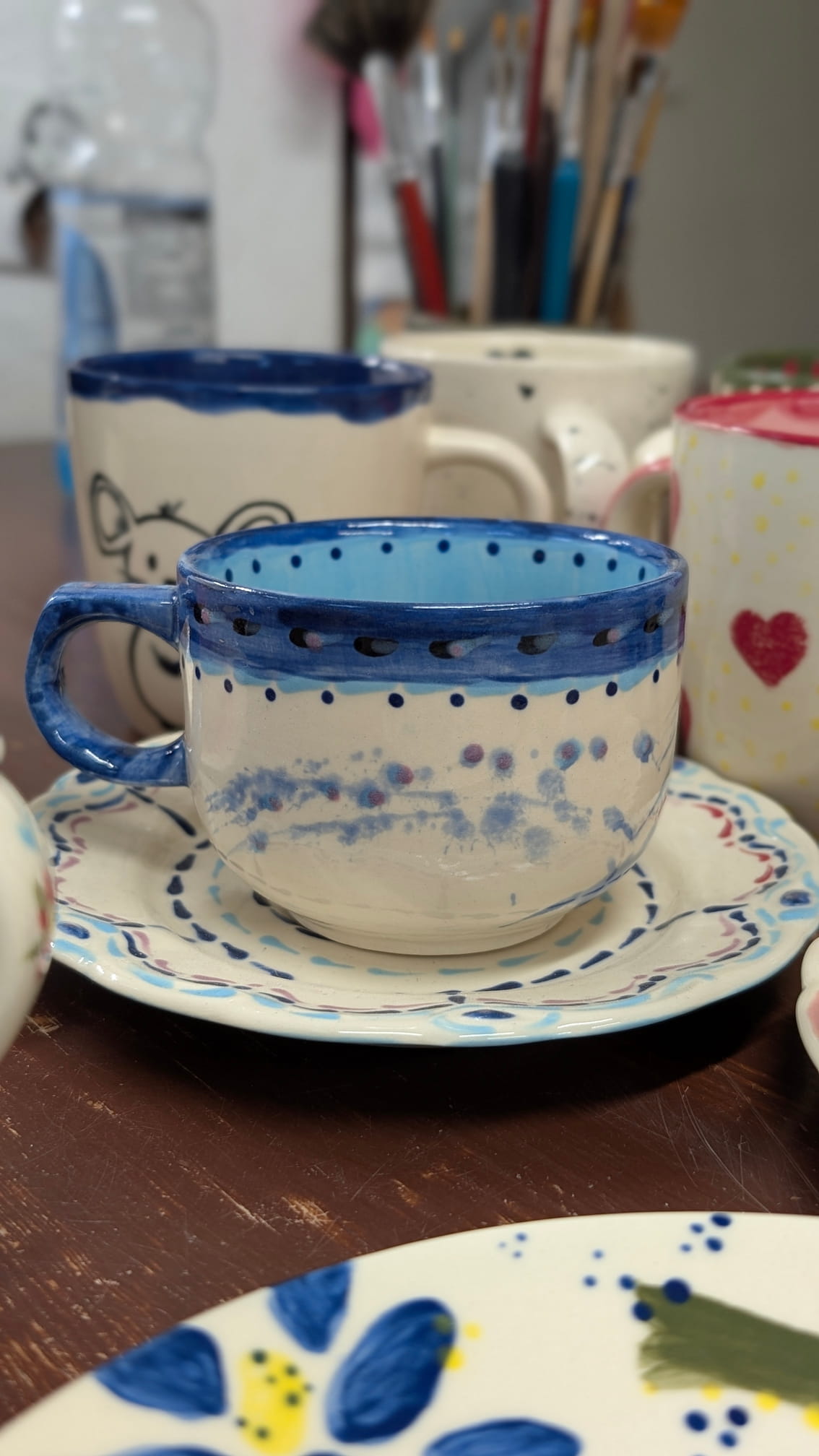 Ceramiczne walentynki 6 Zestaw ceramiczny (filiżanka i spodek) z motywem niebieskich kropek i wzorów. Wnętrze filiżanki jest jasnoniebieskie, a górny brzeg ozdobiony grubym, ciemnoniebieskim pasem z kropkami. Na korpusie i spodku widać rozmyte, akwarelowe plamy w odcieniach niebieskiego i fioletu.